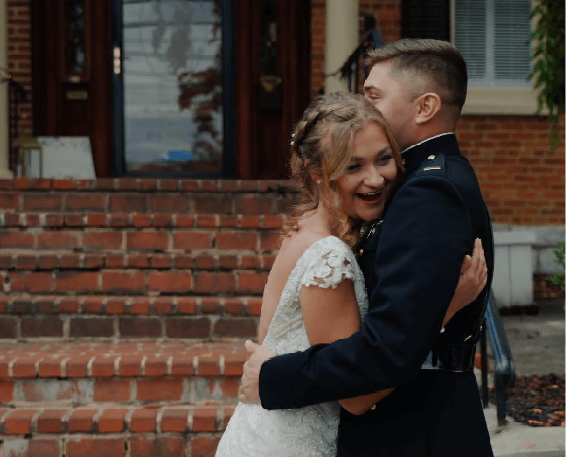 Bride in white lace dress laughing with her groom in uniform on historic brick steps