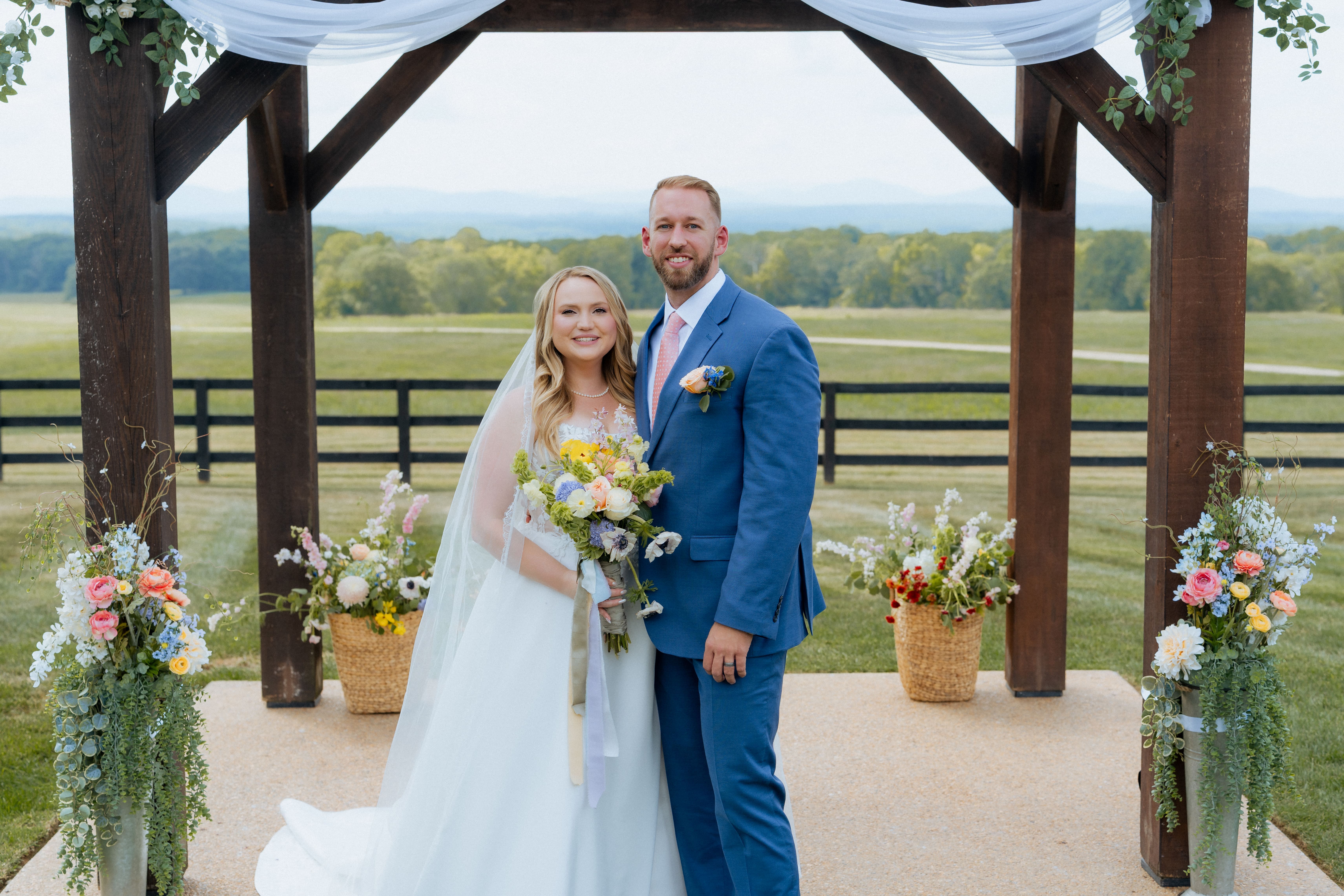 A bride and groom pose together under a wooden gazebo decorated with flowers at an outdoor rural wedding venue with expansive green fields in the background.