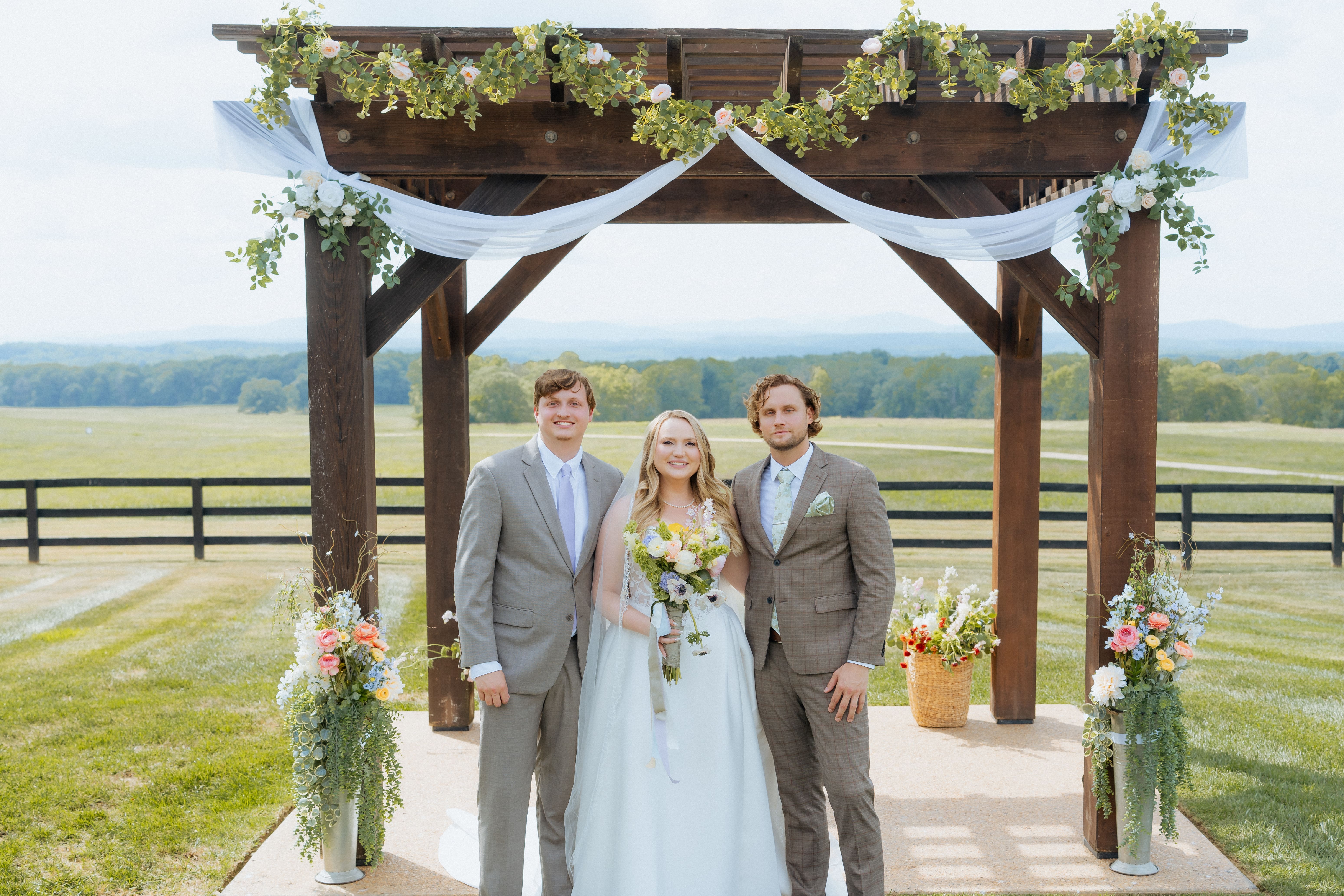 A bride and two men stand beneath a decorated wooden arbor at an outdoor wedding venue with rolling pastures and fencing visible in the background.