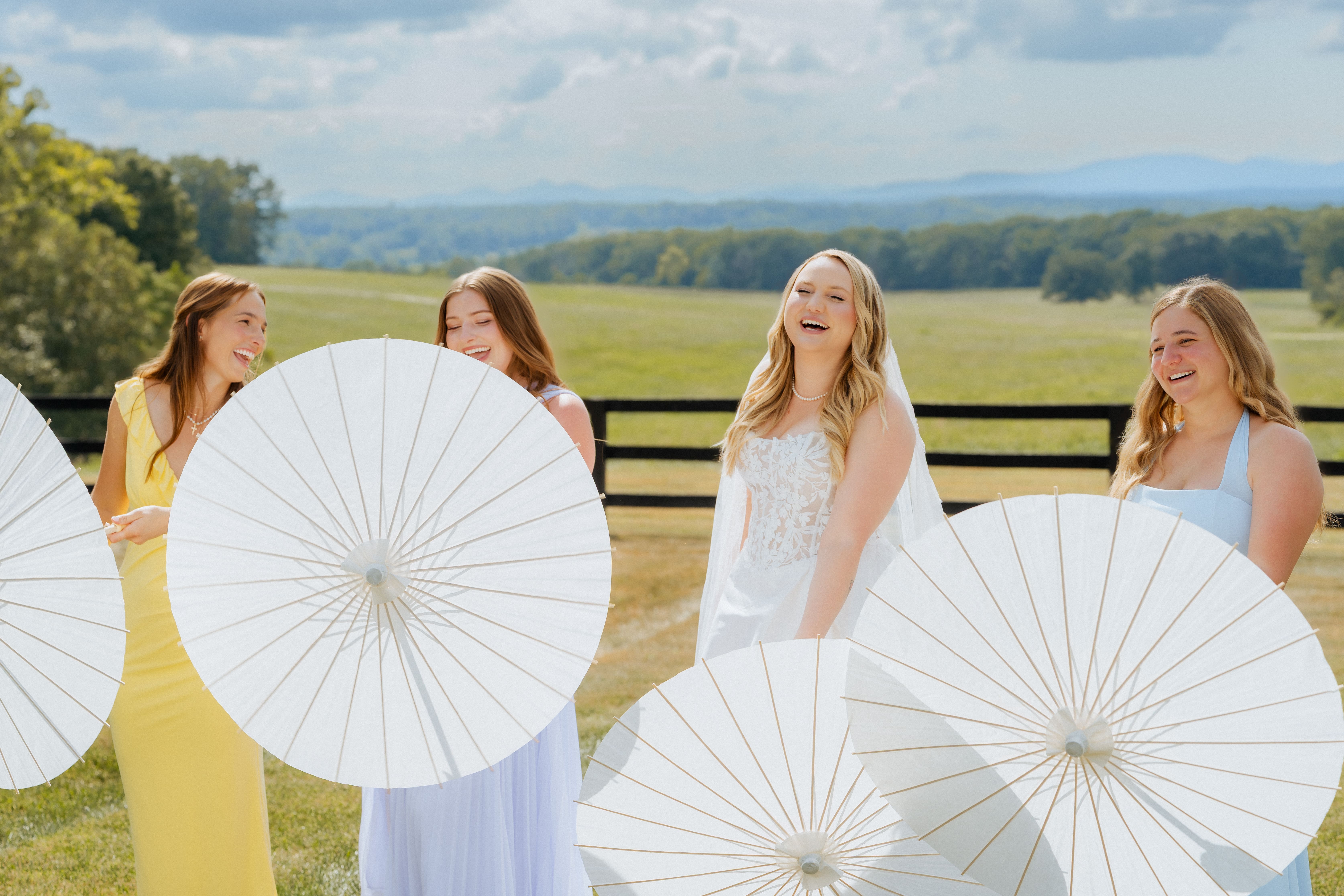 Four young women in white dresses hold white parasols while standing in a sunny countryside setting with rolling green hills in the background.