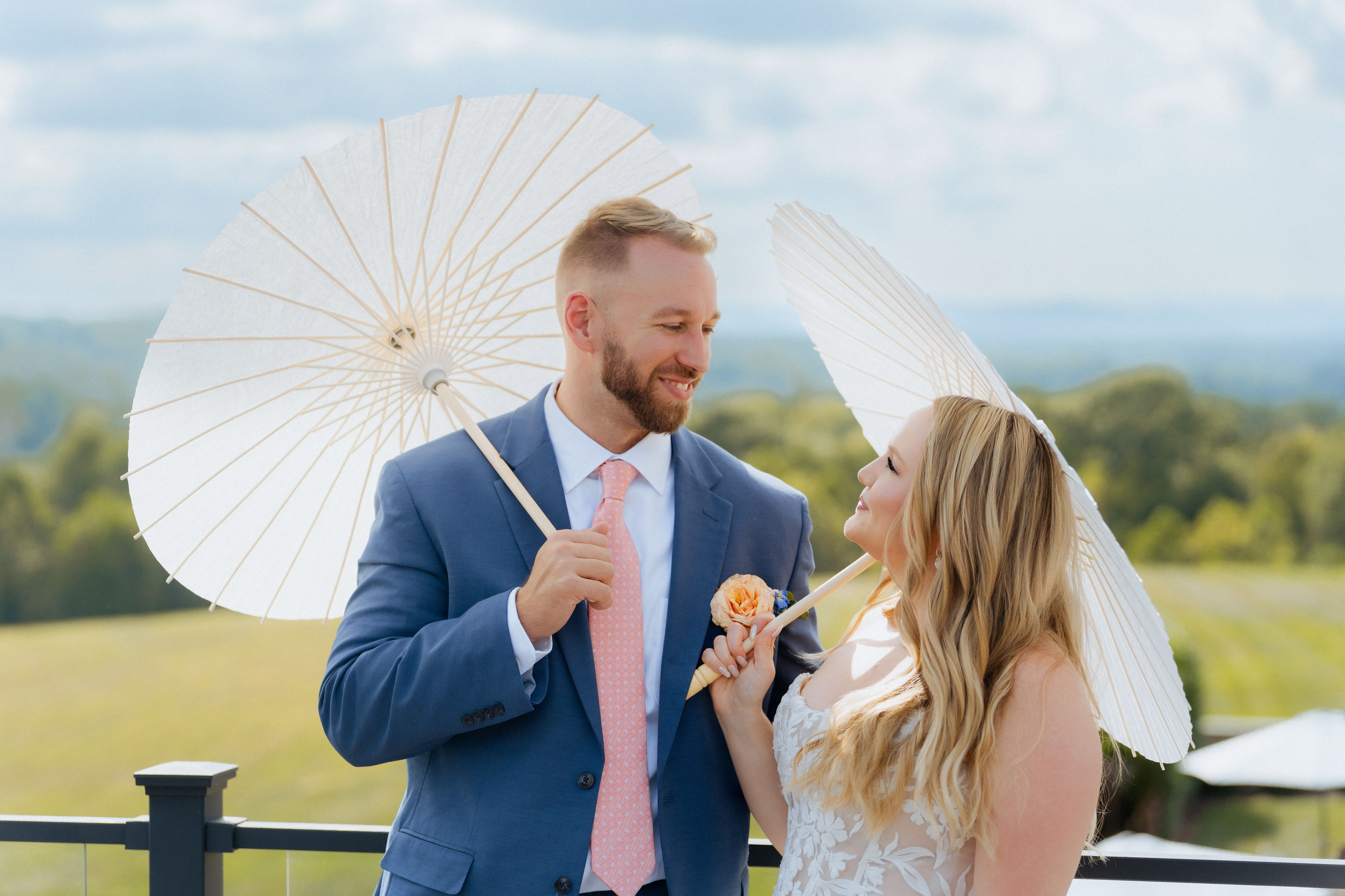 A couple dressed in formal wedding attire holds white parasols while posing outdoors with rolling countryside hills visible in the background.