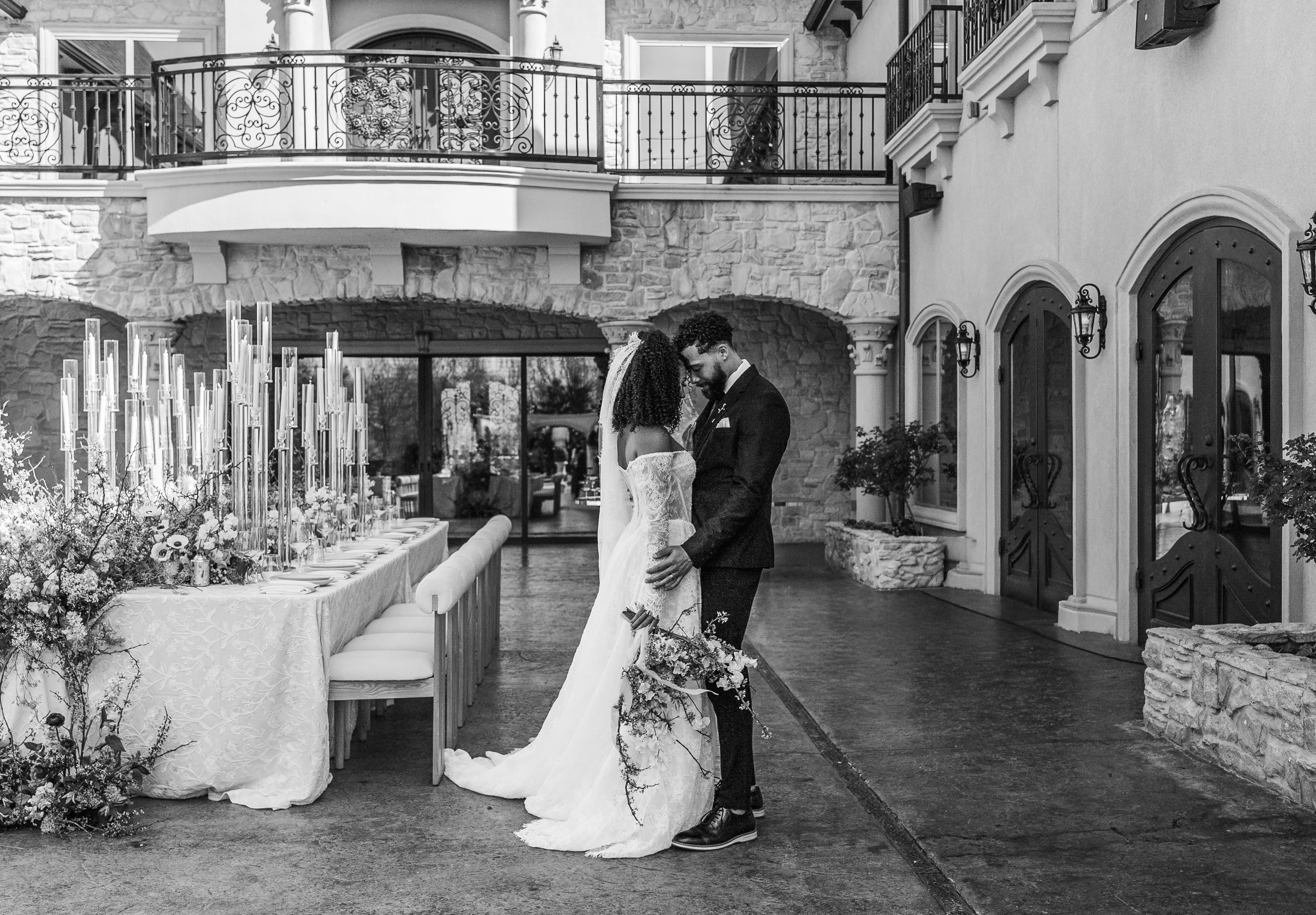 A bride and groom stand together in a Mediterranean-style courtyard venue during their wedding ceremony, with white floral arrangements and architectural arches visible in the background.