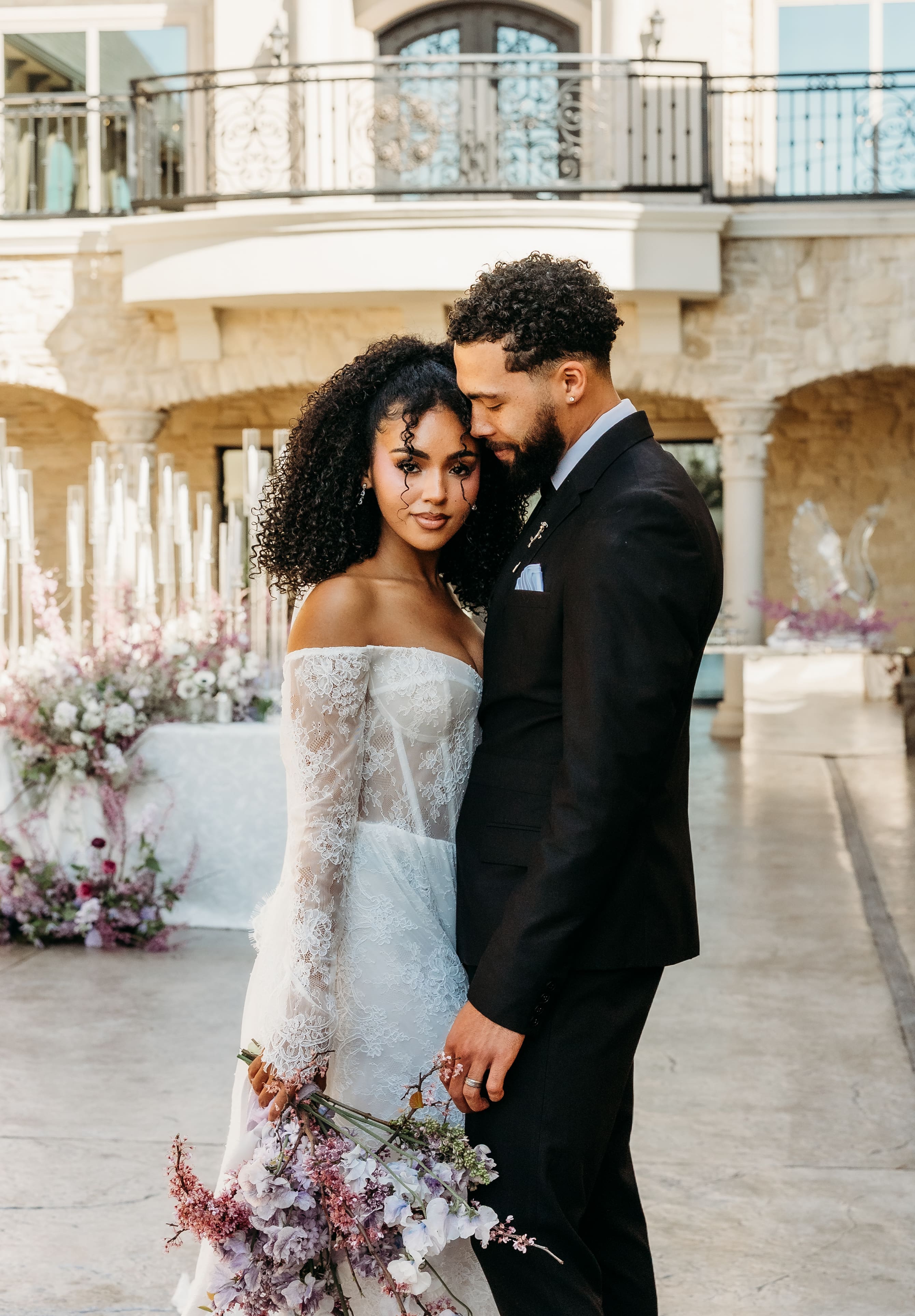 A bride and groom pose together in an intimate portrait at a Mediterranean-style venue during golden hour, with flowering arrangements and stone architecture in the background.