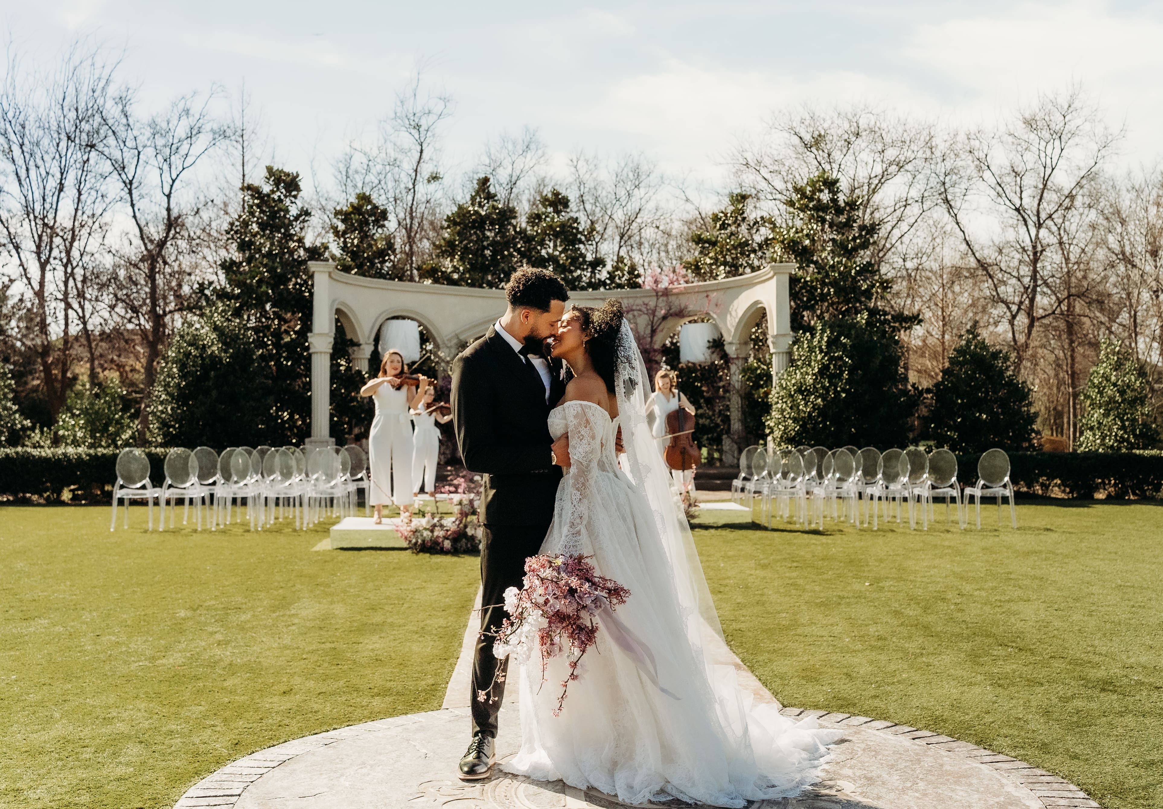 A bride and groom stand together on a lawn in front of a white wedding arch with guests seated in white chairs at an outdoor ceremony venue.