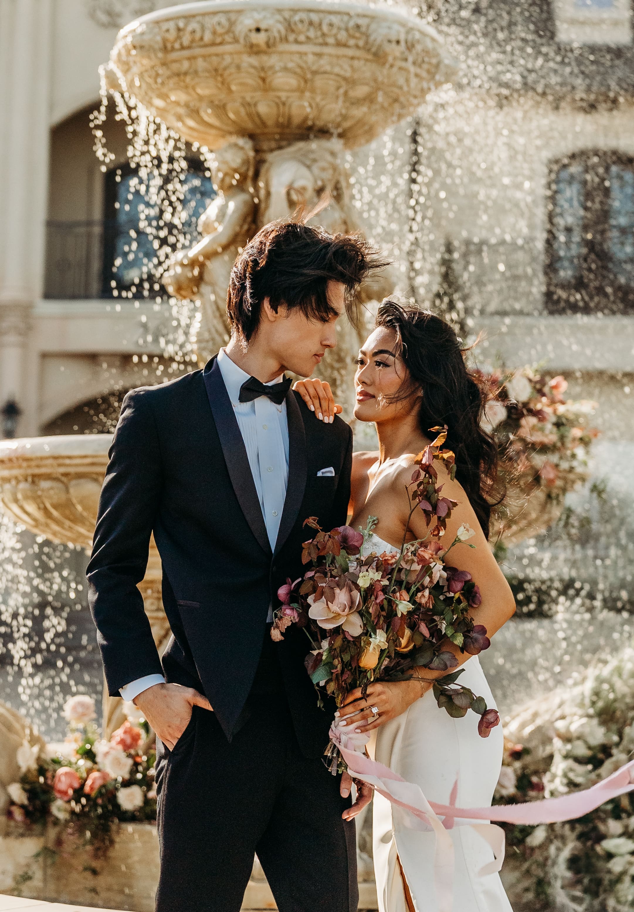 A elegantly dressed couple shares an intimate moment under a golden chandelier with confetti falling around them in a celebratory wedding atmosphere.