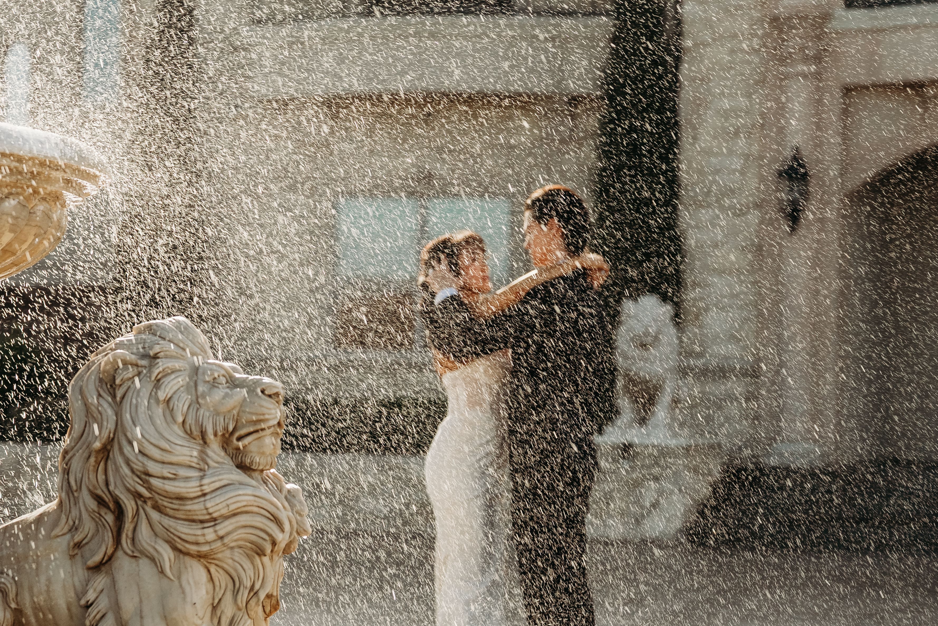 A bride in a white wedding dress holds a bouquet while posing by a classical fountain with water spray in an elegant stone venue.