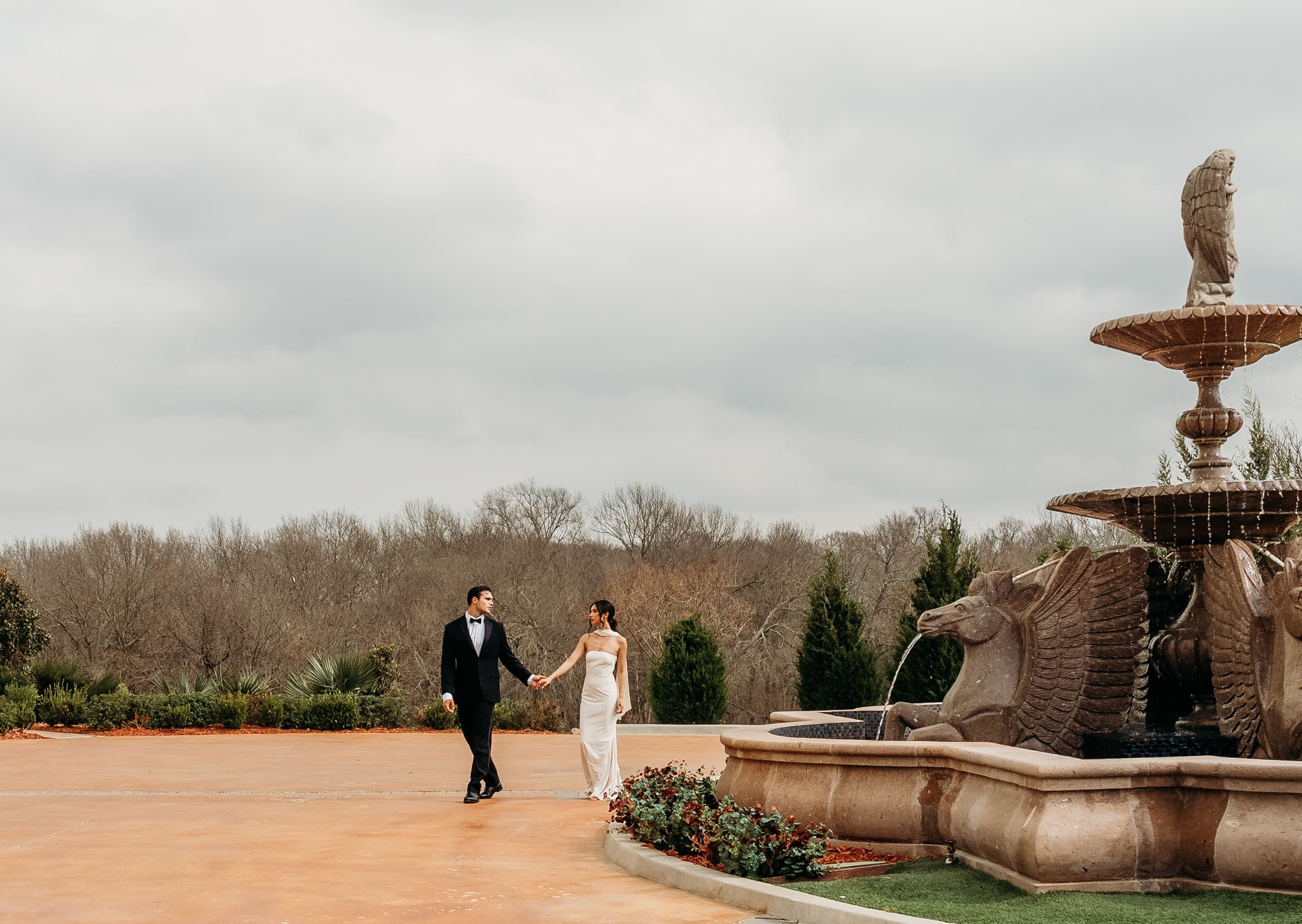 A couple walks near an ornamental stone fountain in a formal garden setting on an overcast day.