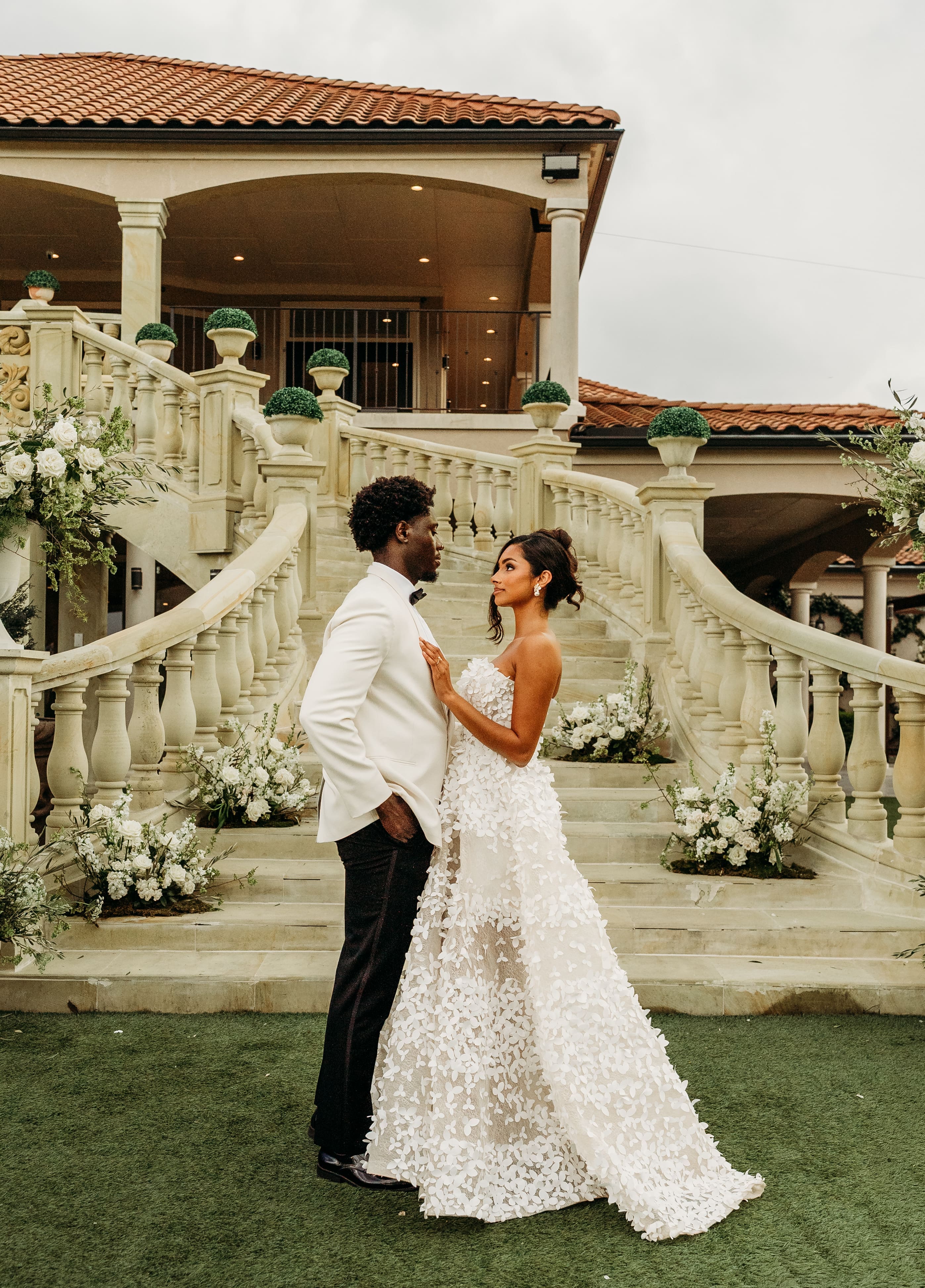 A bride and groom embrace on a manicured lawn in front of an elegant stone mansion with ornate staircases and white floral decorations during their wedding celebration.