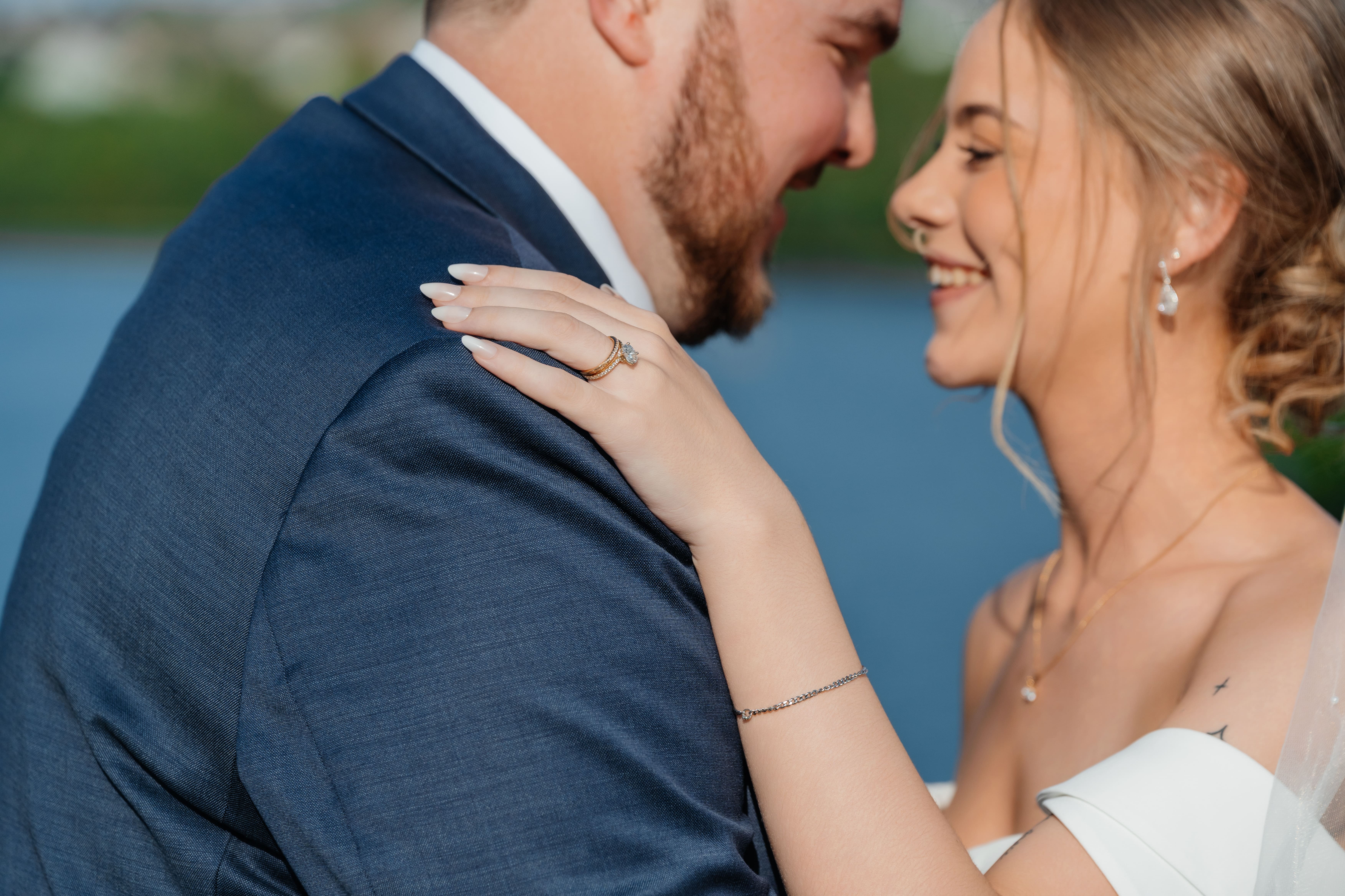 A bride and groom share an intimate moment during their wedding day, posed against a scenic water backdrop.