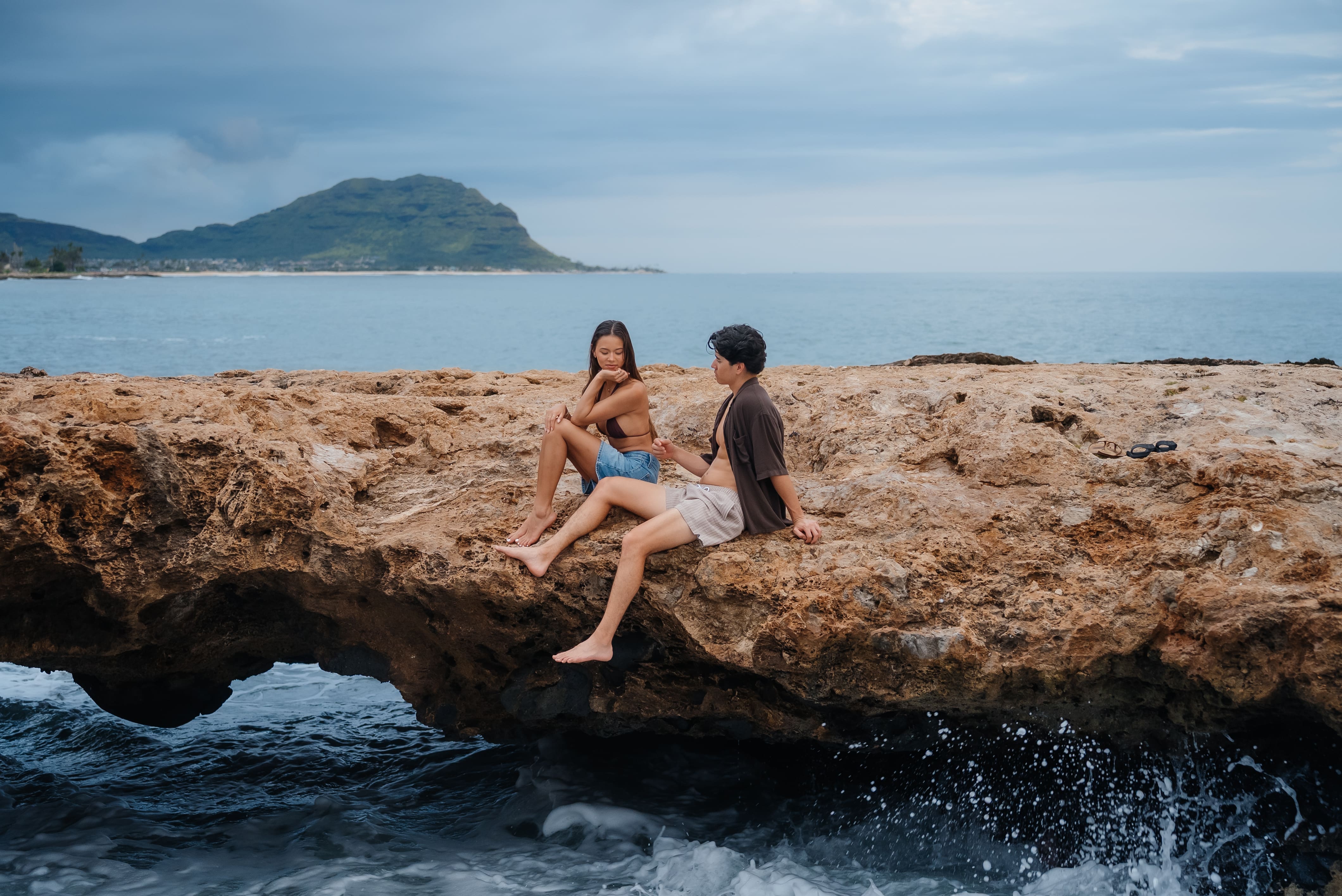 Two young people sit together on a rocky outcrop along a scenic coastal beach with calm blue waters and a distant island in the background.