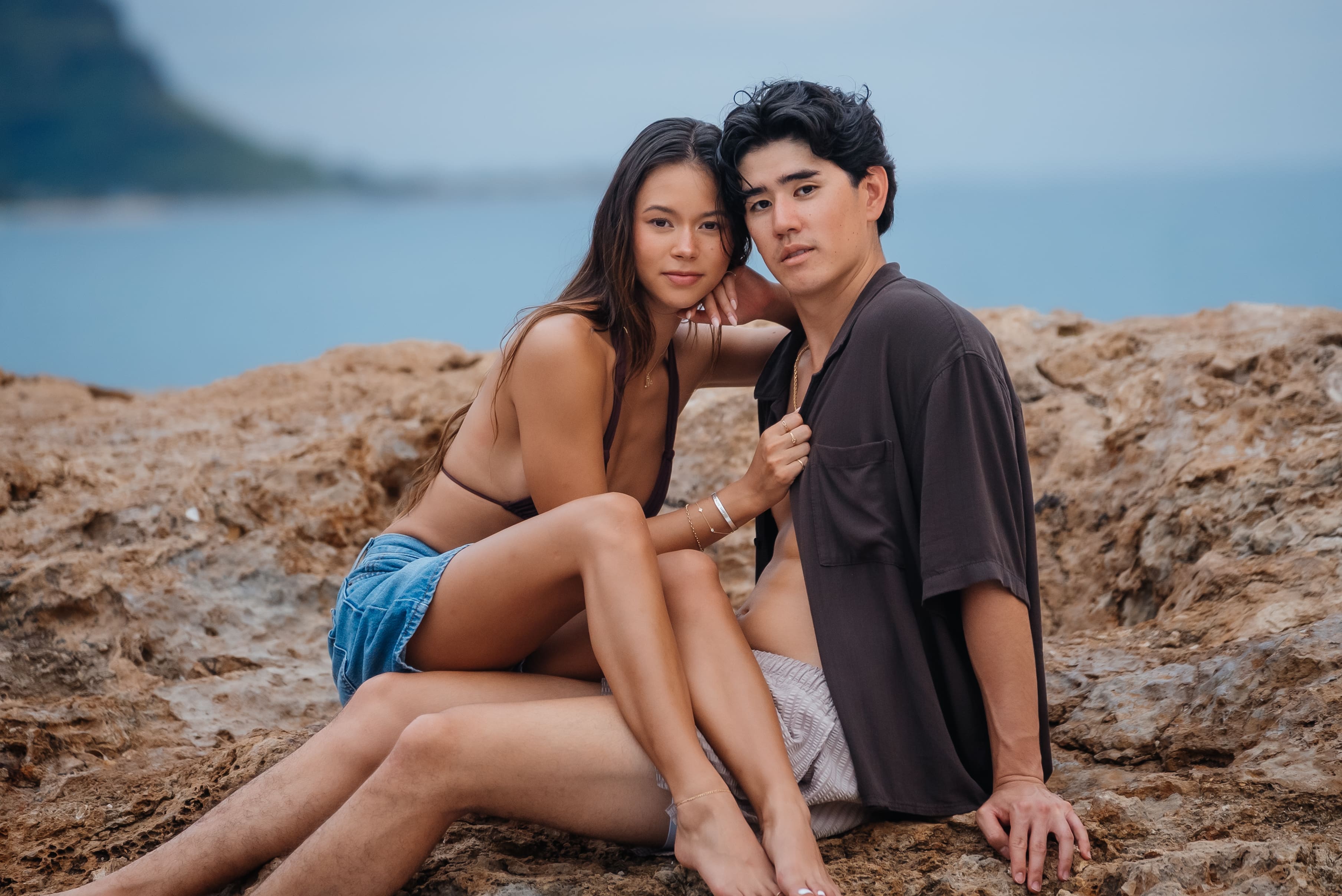 A young couple sits together on a rocky beach during golden hour with calm ocean water in the background.