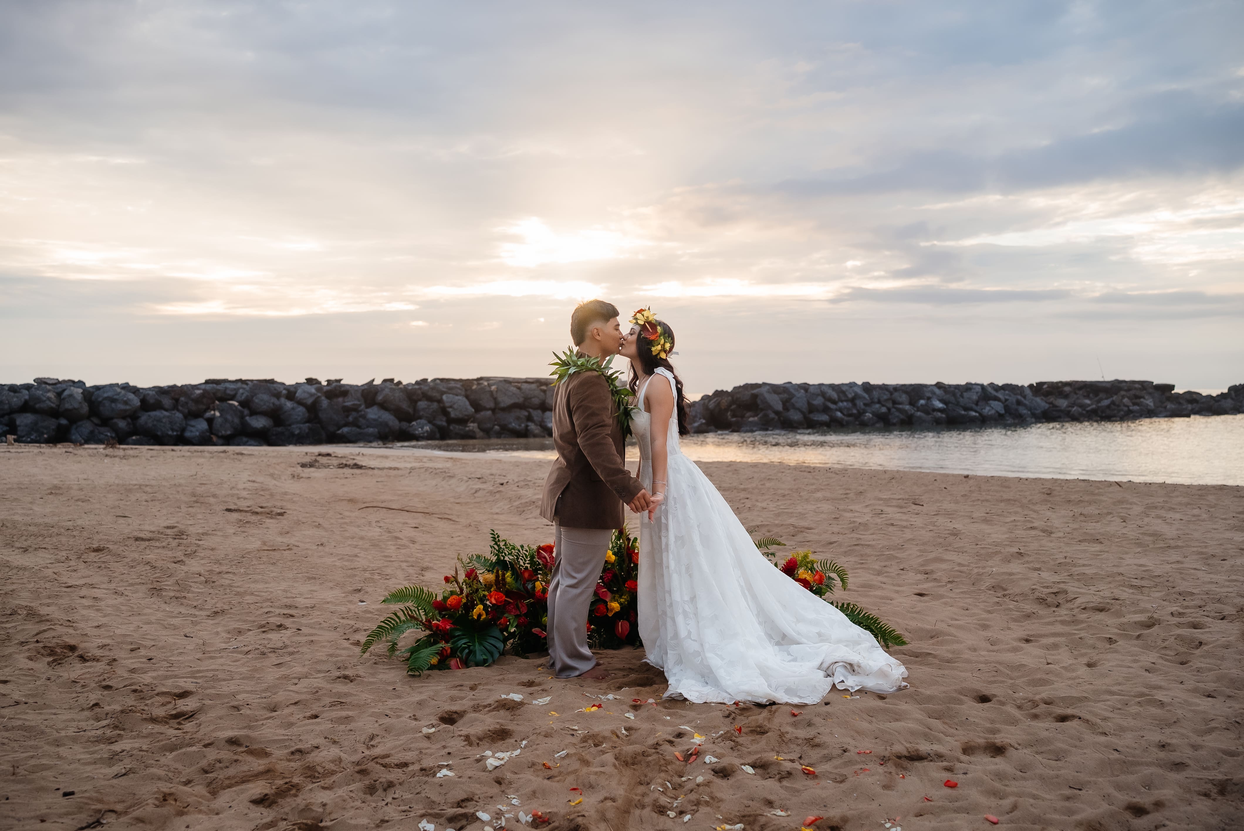 A bride and groom embrace on a sandy beach during golden hour with calm water and floral decor visible in the foreground.