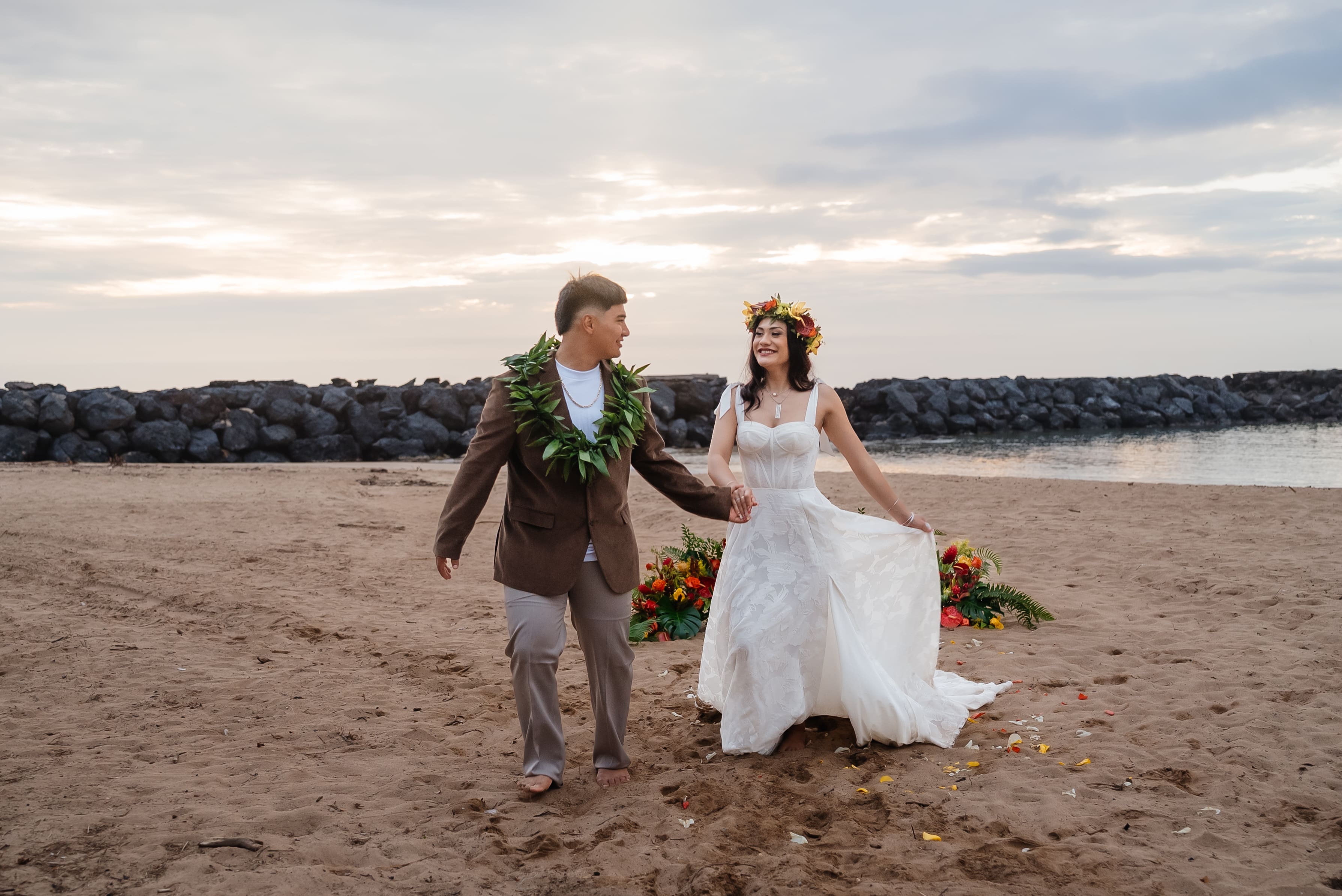 A bride and groom walk together on a sandy beach during golden hour, holding bouquets in a romantic wedding portrait.