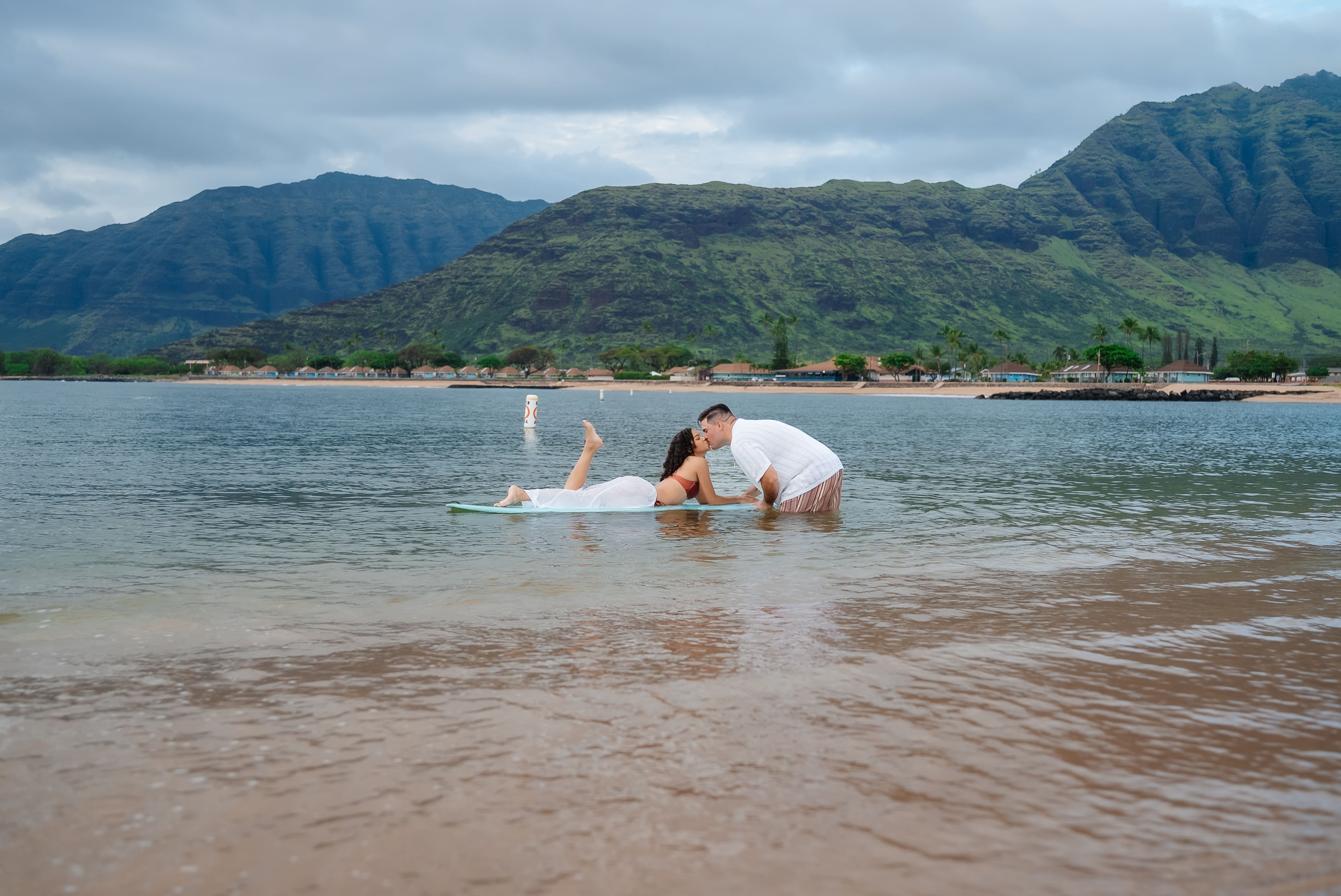 A surfer in a wetsuit rides a white surfboard in calm turquoise ocean waters with lush green mountains visible in the background on a cloudy day.