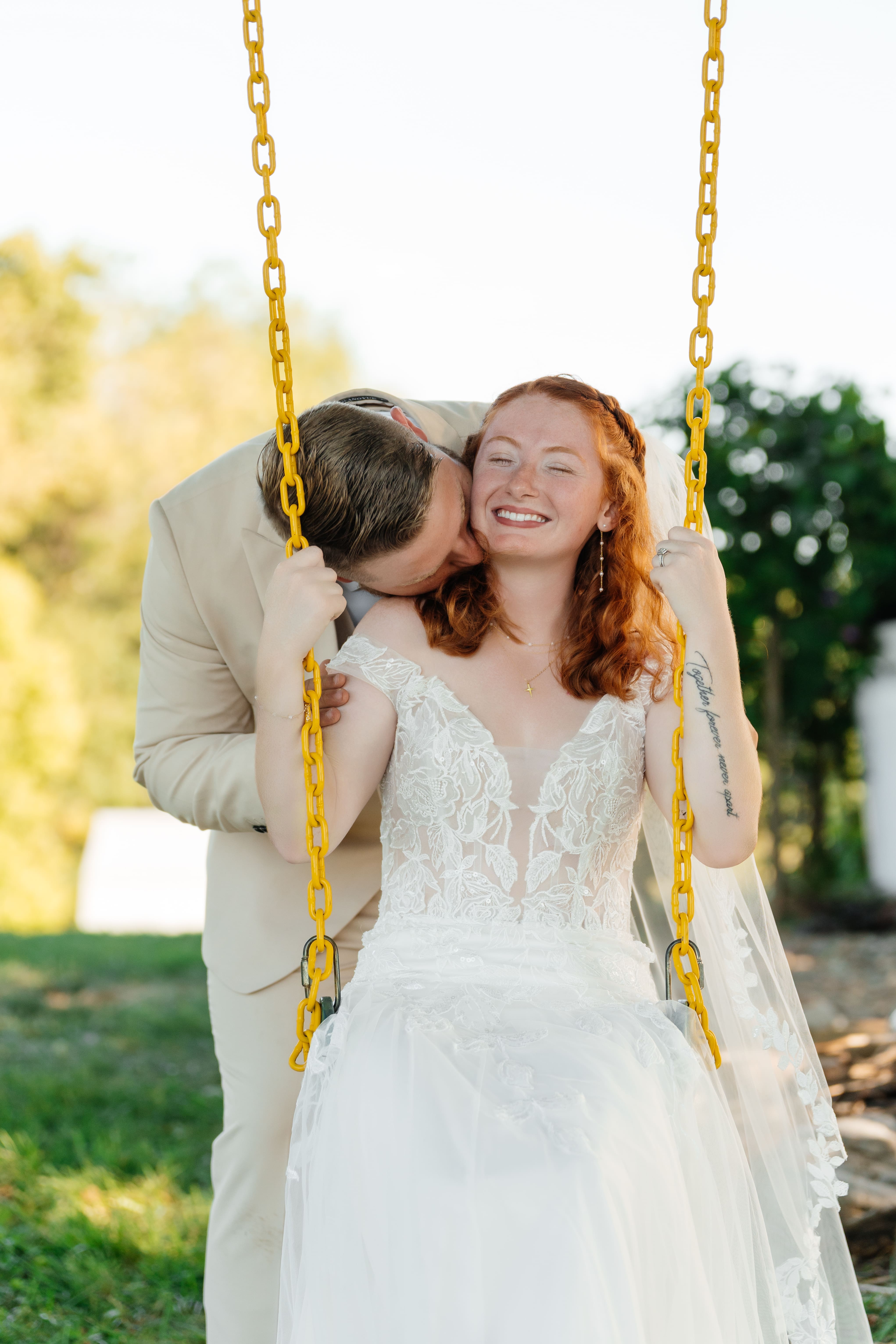 A groom kisses his bride on the cheek while she smiles joyfully on a swing in a sunlit garden during golden hour.