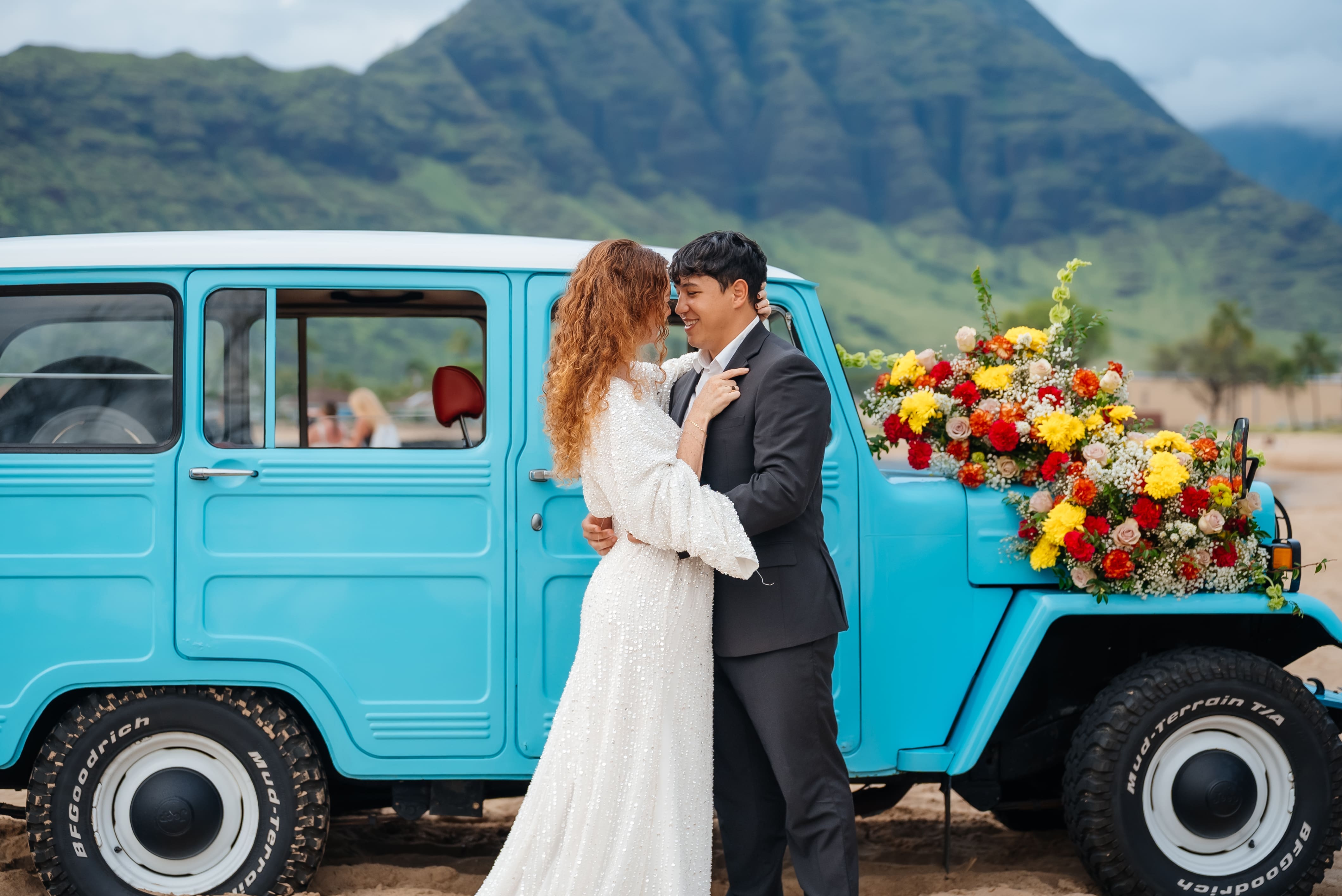 A bride and groom kiss beside a vintage turquoise truck decorated with colorful flowers, with dramatic mountains in the background.
