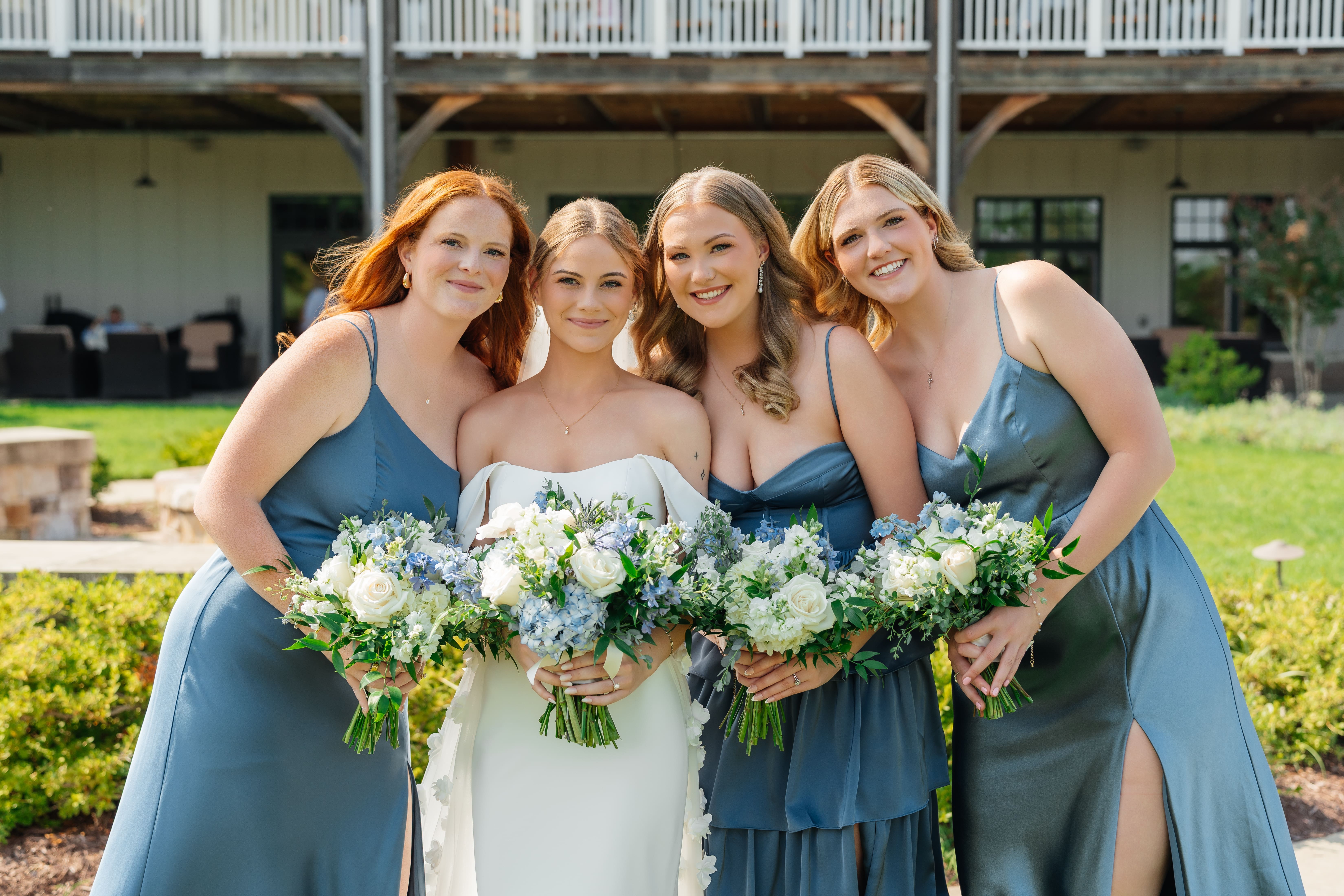 A bride and her bridesmaids pose together outdoors holding white and greenery bouquets in front of a colonial-style venue with manicured lawn.