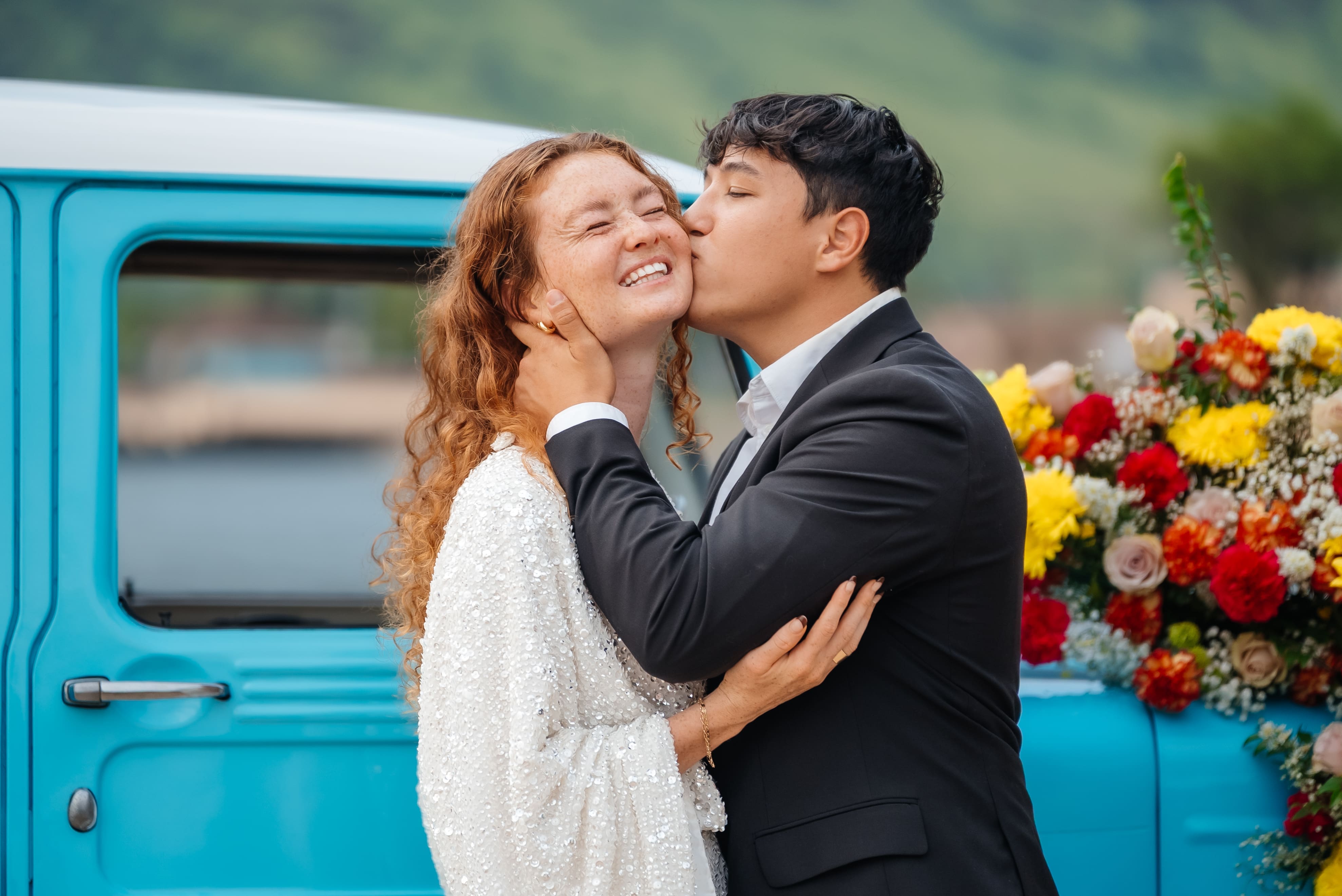 A newlywed couple shares a romantic kiss in front of a vintage turquoise truck decorated with colorful flowers.