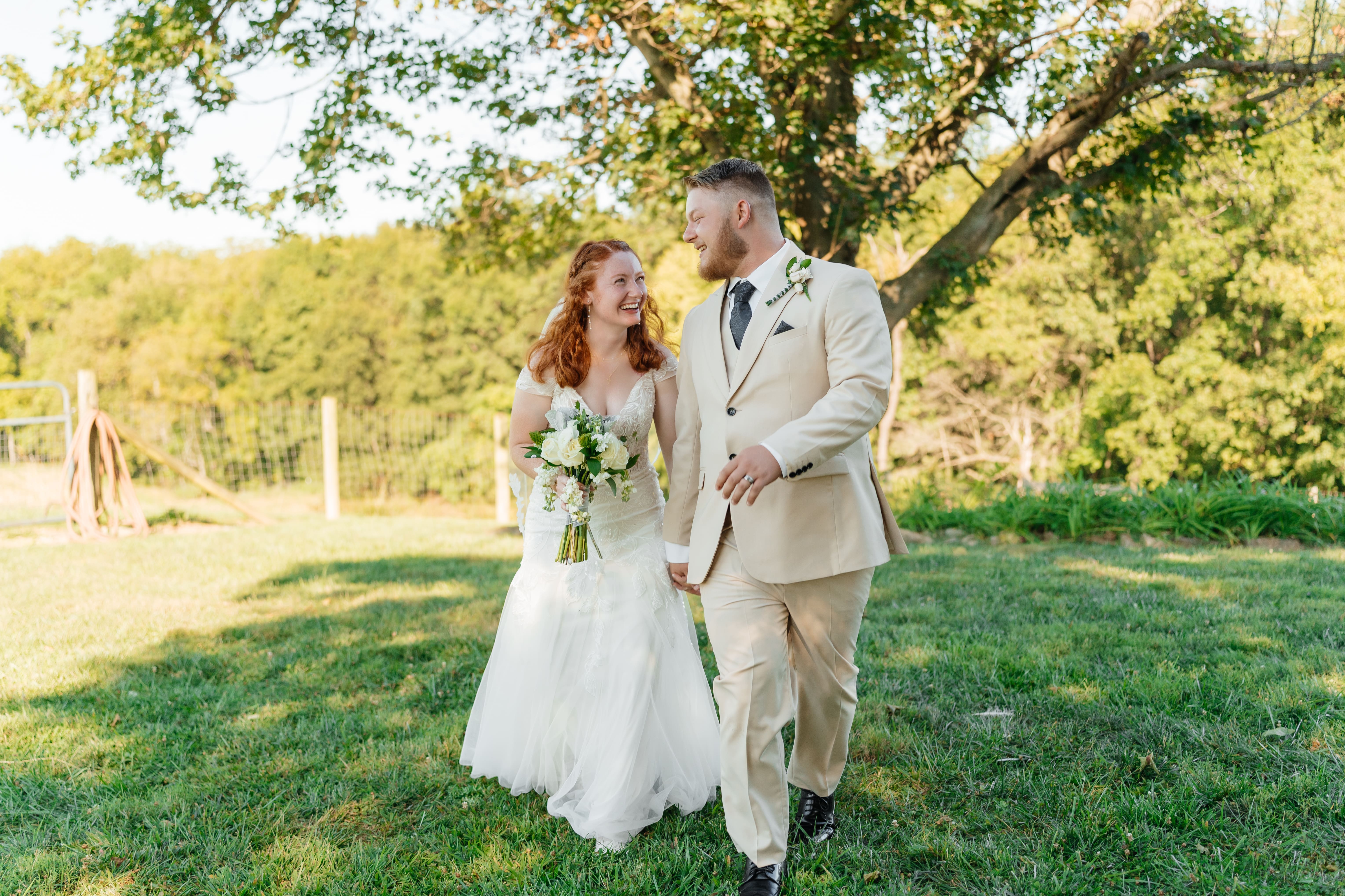 A bride and groom stand together outdoors in golden hour light, with the bride holding a bouquet and both smiling at the camera in a natural park-like setting.