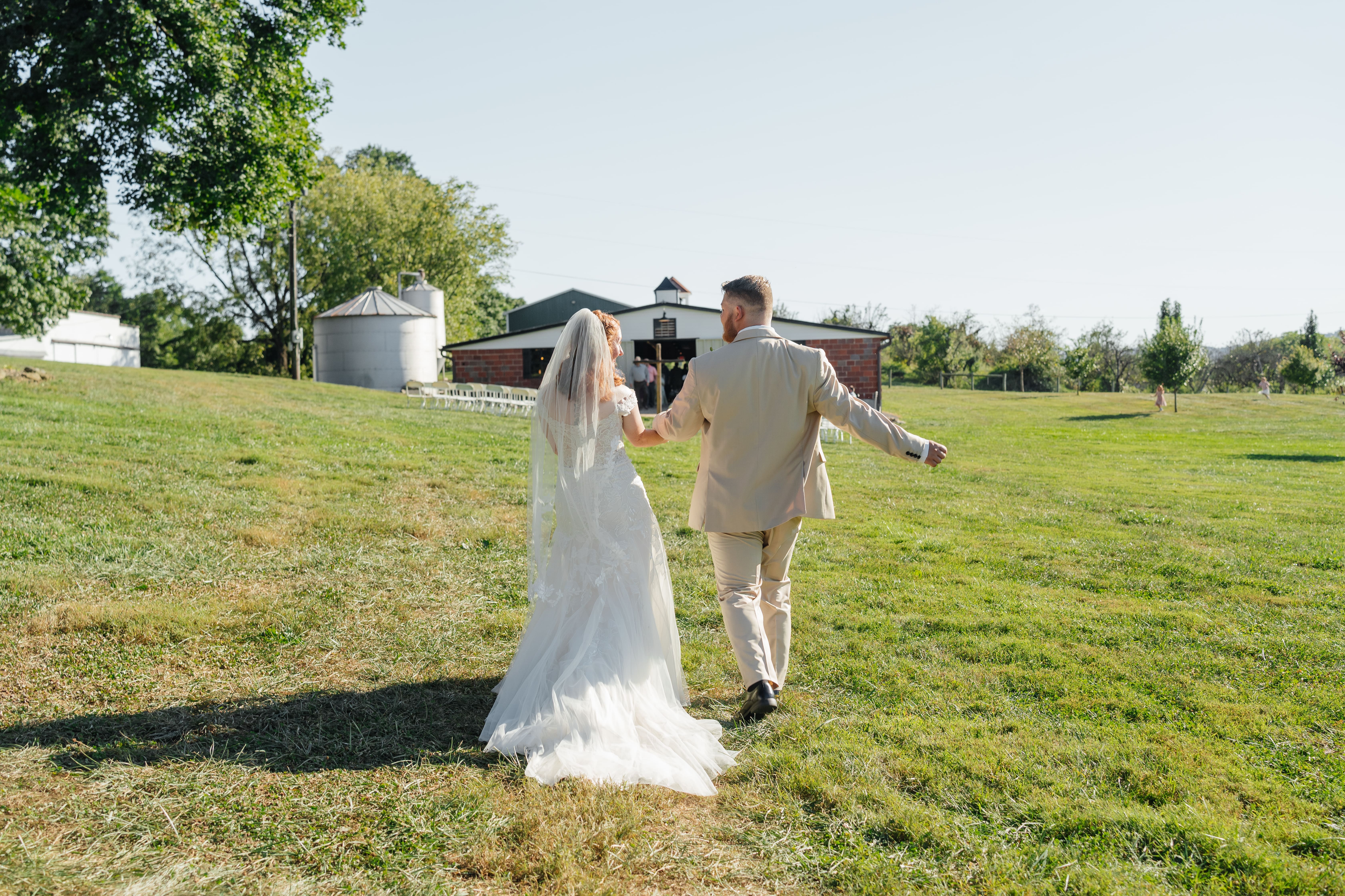 A bride and groom walk hand-in-hand through a spacious green pasture on their wedding day, with a barn visible in the background.