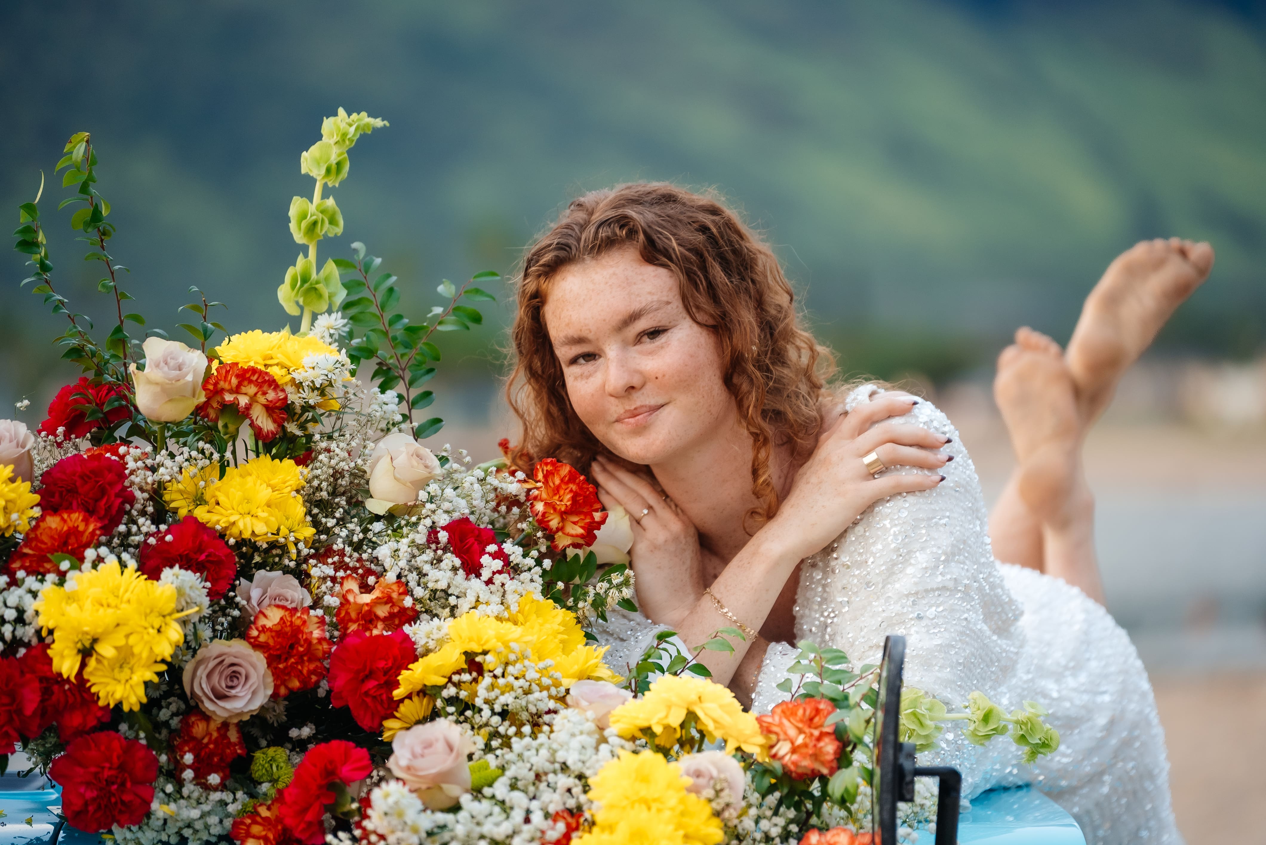 A smiling woman with reddish-brown hair poses beside a vibrant bouquet of red, yellow, and white flowers against a blurred outdoor background.