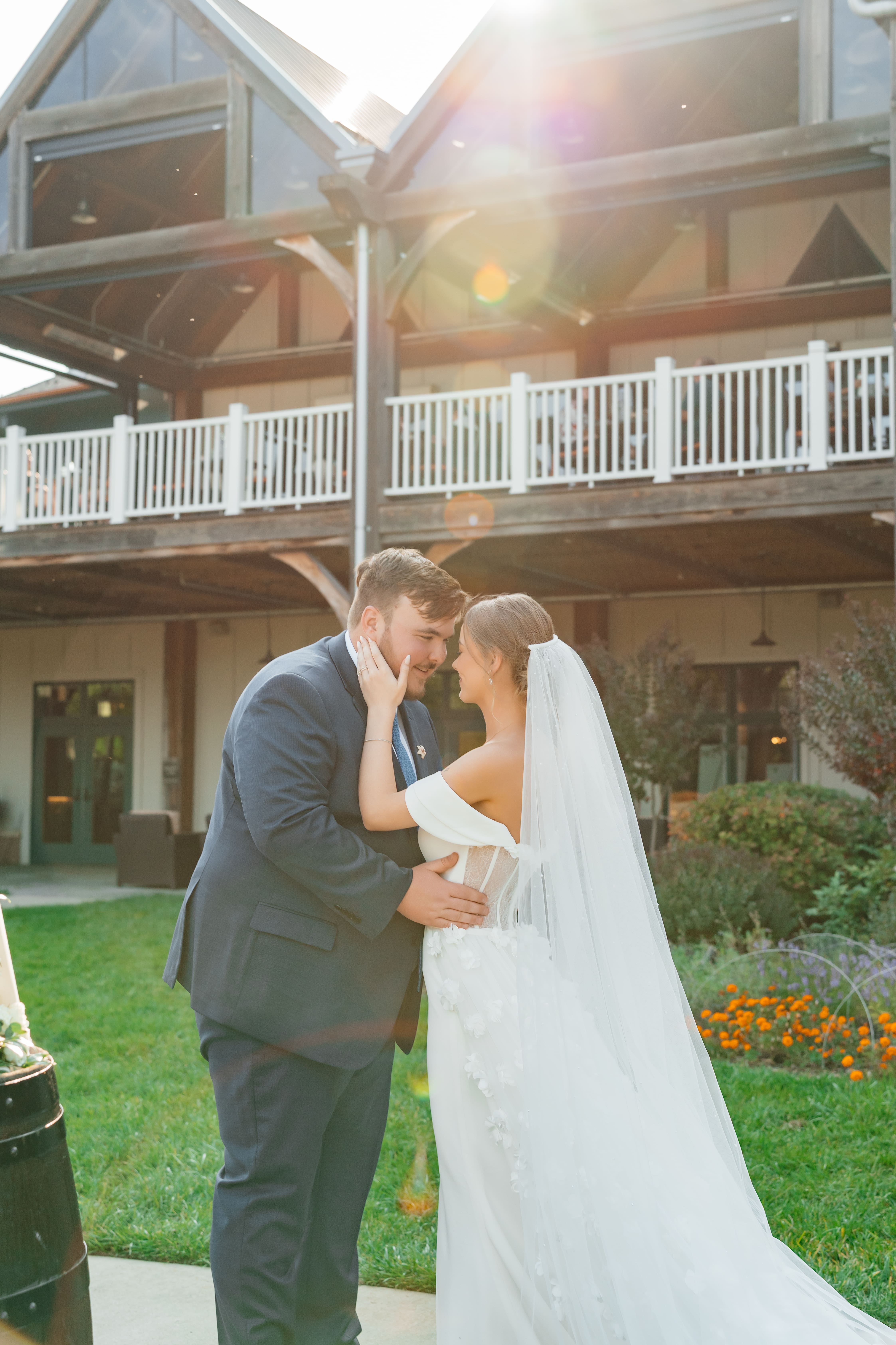 A bride and groom share a kiss during their wedding ceremony in front of an elegant mansion with white railings, backlit by golden hour sunlight.