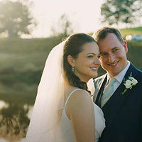 A bride and groom pose together outdoors during golden hour, with the bride wearing a veil and the groom in a dark suit with a white boutonniere, set against a softly blurred garden background.