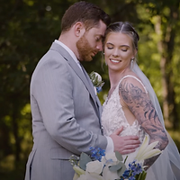 A bride and groom pose together outdoors during golden hour, with the bride displaying a tattoo on her arm while holding a floral bouquet.