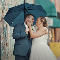 A bride and groom pose together under a black umbrella against a weathered brick wall in an intimate rainy wedding portrait.
