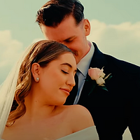 A newlywed couple poses together outdoors during golden hour, with the bride wearing a veil and the groom in a dark suit with a boutonniere, set against a soft sky backdrop.