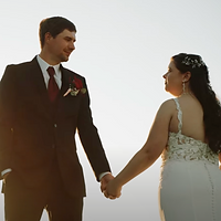 A bride and groom hold hands while posing for a portrait against a soft, minimalist background during golden hour lighting.