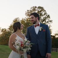 A bride and groom stand together outdoors during golden hour, the bride holding a white floral bouquet while gazing at her groom in a serene countryside setting.