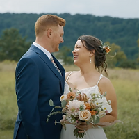 A bride and groom pose together in a pastoral field during golden hour, with the groom looking at the bride who holds a bouquet of flowers.
