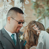 A bride and groom share an intimate moment during their wedding ceremony, with the bride wearing a delicate lace veil and floral crown while the groom wears glasses and a gray suit with boutonniere.