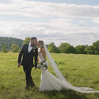 A bride and groom walk through an open meadow on their wedding day, with the bride's flowing veil trailing behind them under a partly cloudy sky.