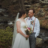 A bride and groom stand together outdoors among moss-covered rocks during golden hour, with the bride holding a bouquet and both dressed formally for their wedding.