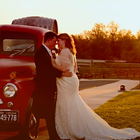 A bride in a white wedding dress poses against a vintage red truck during golden hour in a rural countryside setting.