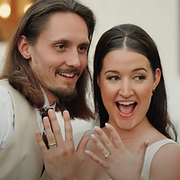 A couple joyfully displays an engagement ring with excited expressions in an indoor setting.
