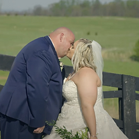 A bride and groom share an intimate kiss in a pastoral outdoor setting with a green field and wooden fence in the background.