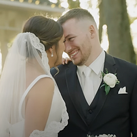 A bride and groom share an intimate moment together outdoors during golden hour, with the bride adjusting her veil near the groom's face in a joyful, tender gesture.